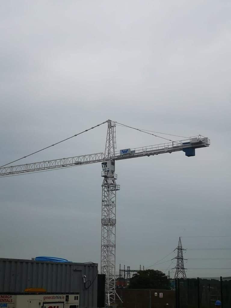 A crane is hoisting a public utility generator to a construction site outdoors, surrounded by a cloudy sky.